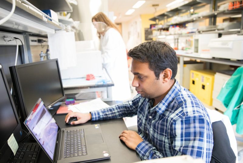 Barath, in a blue plaid shirt, sits in front of his laptop with research results on the screen, and the background is the lab setting.