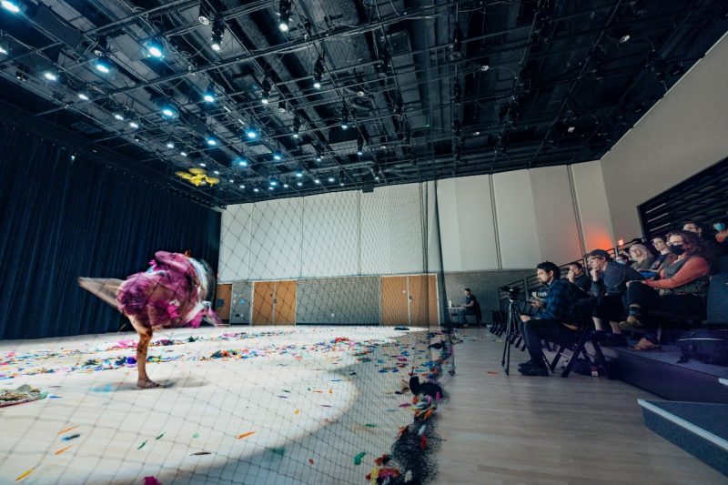 Behind a safety net, dancer Scotty Hardwig moves in a blur alongside a yellow drone. An audience in the Creativity and Innovation District watches.