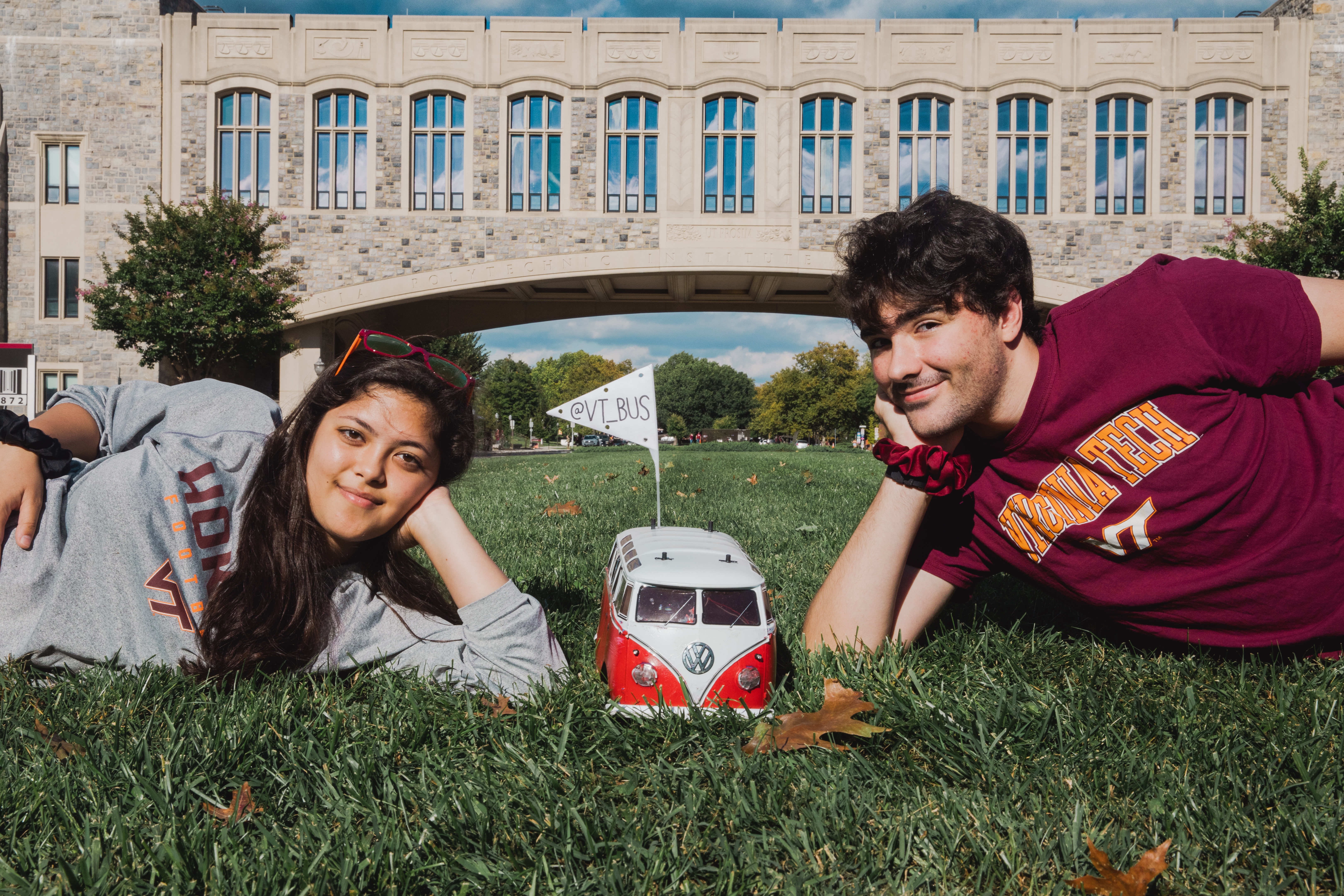 Two students lay in the grass with their heads in their hands, beside their remote-controlled Volkswagen bus. The bus is white and red and has a small flag on top.
