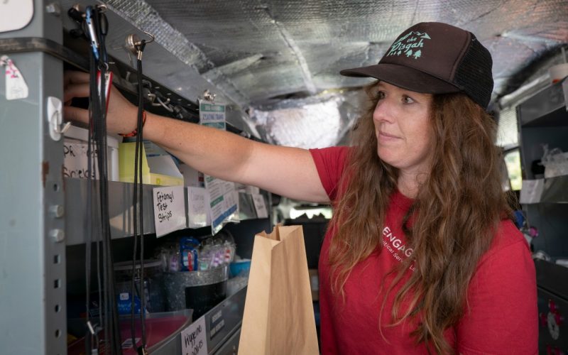 Woman wearing red shirt and ball cap holds a brown paper bag and reaches for medical supplies organized on shelving.