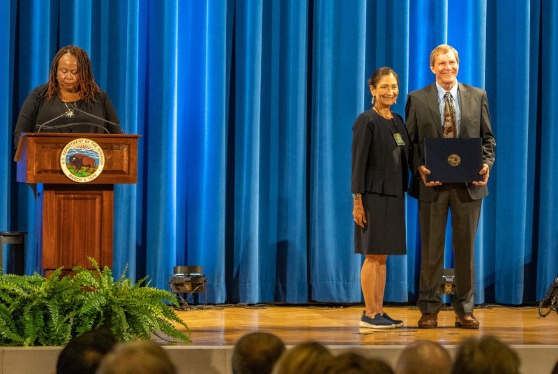 : Secretary of the Interior Deb Haaland stands next to Jeff Marion, who holds an award certificate.  