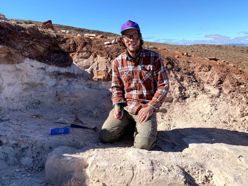 A man, wearing a plaid shirt and baseball cap, kneels and smiles for a photographer as he takes part in a rural fossil dig.
