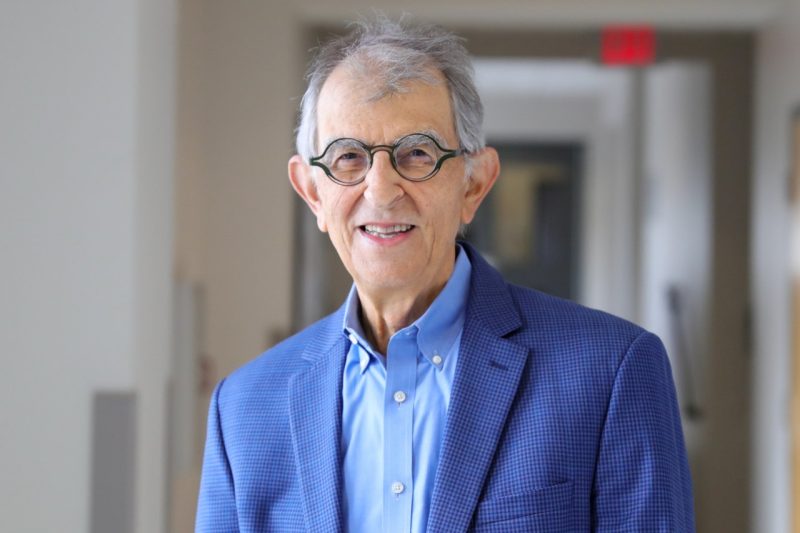 A man with owl-like glasses and a sharp blue sports coat and pale blue shirt poses for a photo in a hallway.