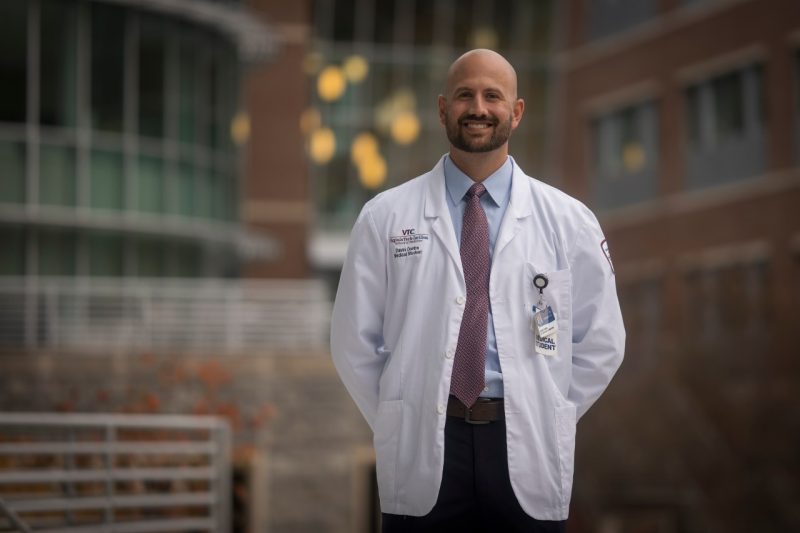 Davin Combs, a fourth-year medical student, stands in his white coat in front of the Virginia Tech Carilion School of Medicine building.