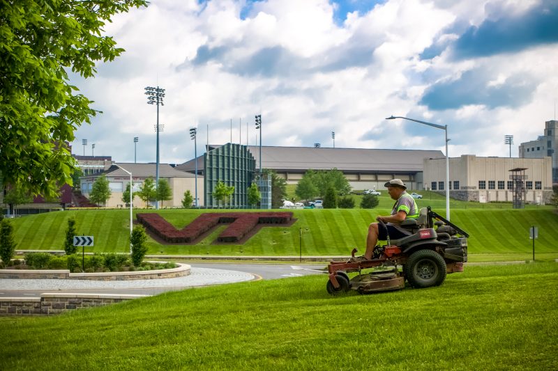 A person drives a lawnmower in the grass on a sunny day. A maroon bush shaped in the letters "VT" sits on a hill in the distance. 