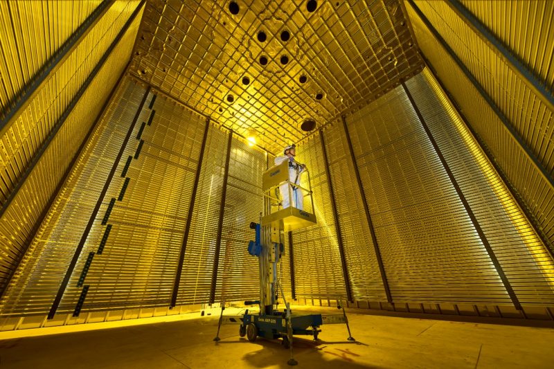A man stands on a lift inside one of the main chambers of the Deep Underground Neutrino Experiment or DUNE for short. The walls of the chamber reflect light.