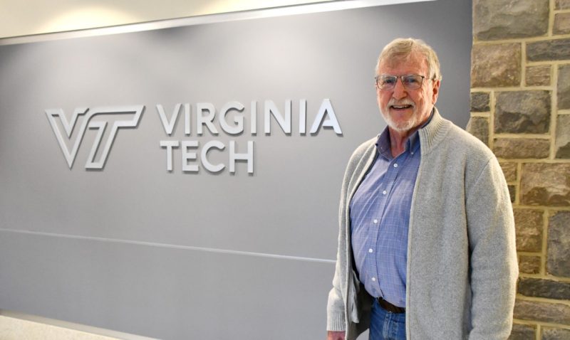 A man wearing an open gray sweater over a blue shirt poses near a Virginia Tech screen.