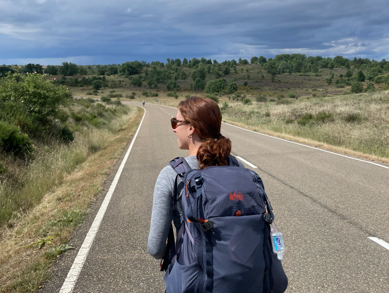 Young woman with backpack faces away from camera looking over a long road that disappears into the horizon. 