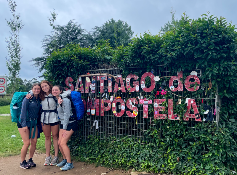 Three female backpackers pose in front of a sign that reads Compostela de Santiago