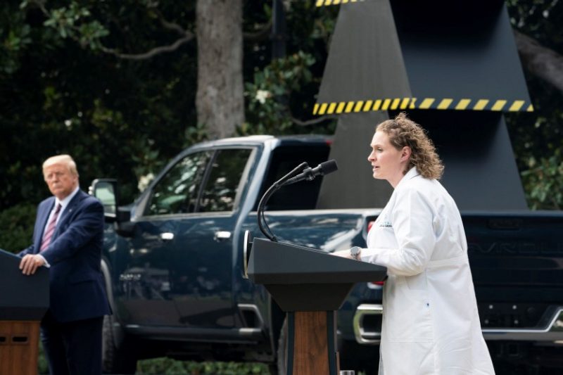Amy Johnson, dressed in her medical whites, speaks at a podium with President Trump and a green pickup truck in the background