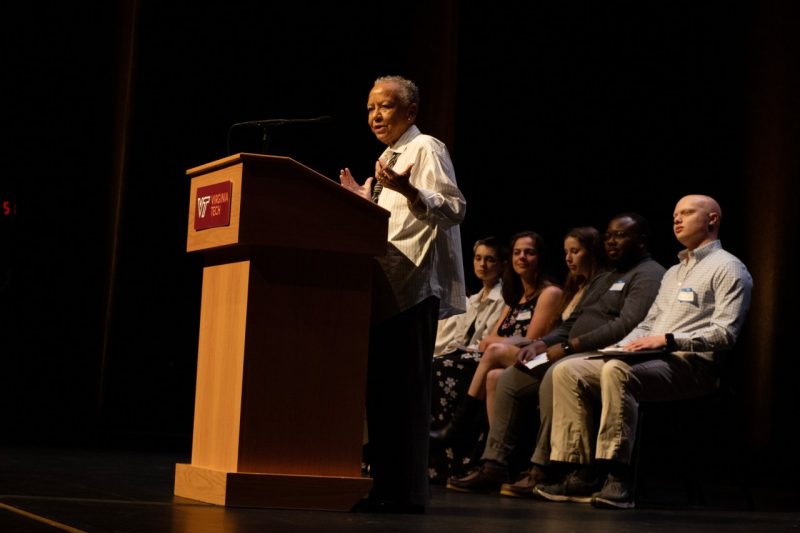 Nikki Giovanni (at left) speaks at a podium with students seated in chairs behind her.