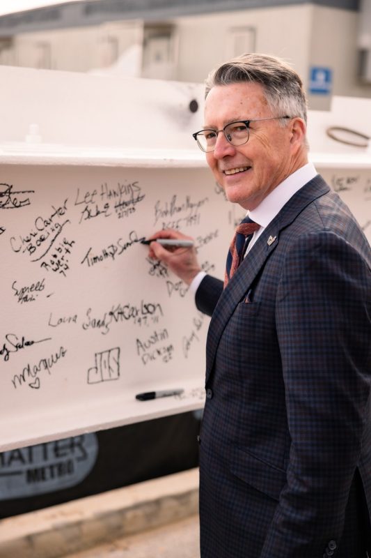A man smiles at the camera while signing his name to a steel beam