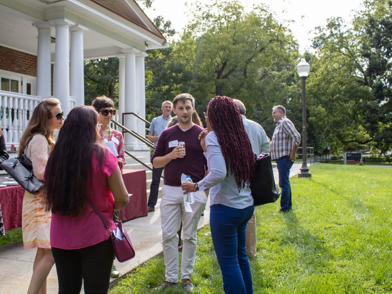 This images shows several faculty standing in small groups conversing in a yard with a table of food options.