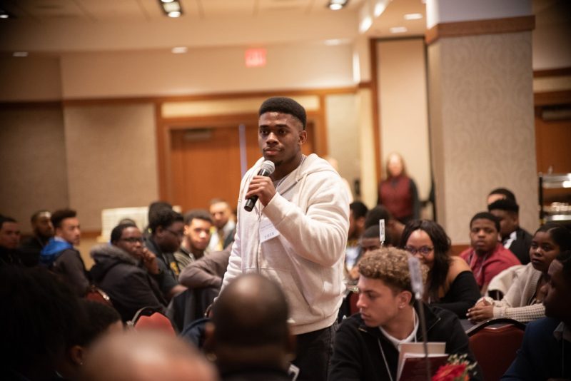 A Black male student in a white sweatshirt holds a microphone during a session at the Uplifting Black Men Conference at the Inn at Virginia Tech.