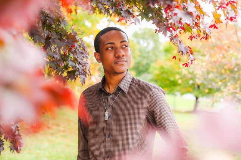 Sherlock Banks, a Black male student in a plaid button-down shirt, is seen smiling through a haze of leaves on campus at Virginia Tech.