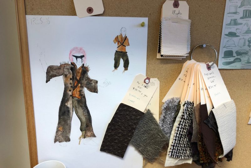 A sketch of a costume for the play "Oedipus" is on a bulletin board, along with labeled fabric swatches in brown and grey shades.