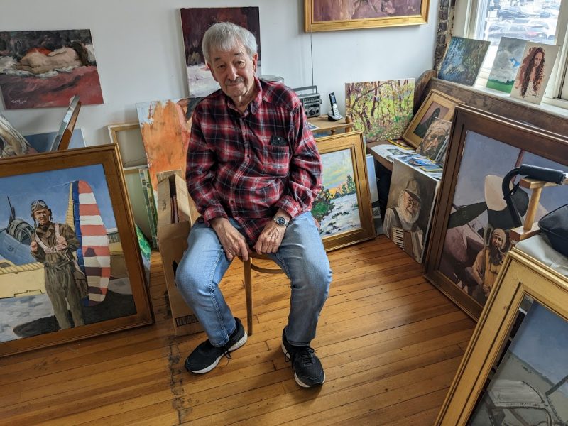 Terry Lyon sits in a chair in his studio. Paintings of his father posing with his World War II aircraft are on either side, and many other paintings are mounted on a red brick wall behind him.