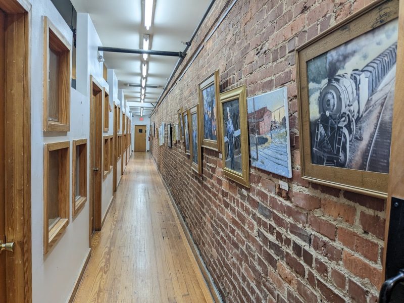 A long hallway, with a red brick wall to the right and wood-framed entrances to artist studios on the left. Several of Terry Lyon's paintings depicting trains and railroad workers are mounted on the wall to the right.