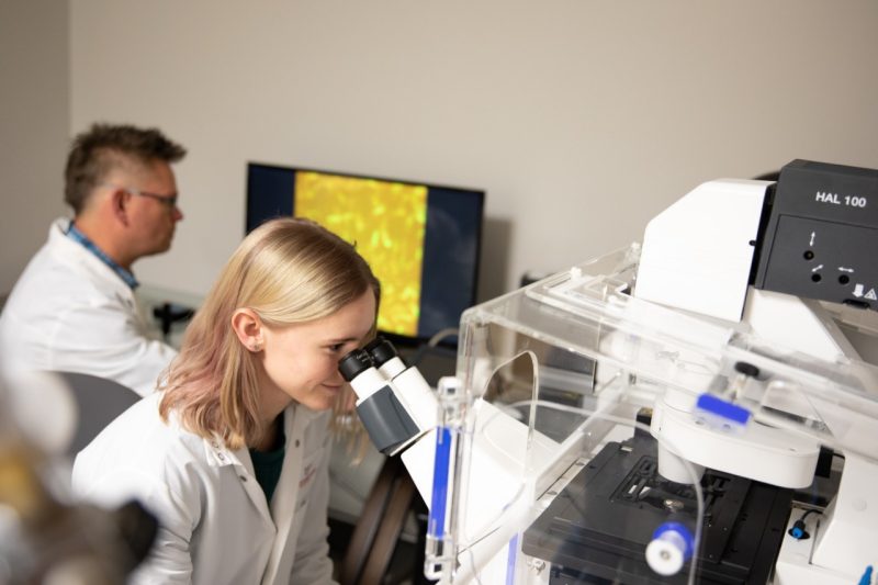 Researchers in white coats look into a microscope.