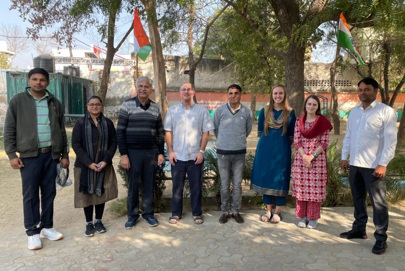 Eight people stand in a semi-circle on the grounds of a rural school, with two flags of India raised in the background.