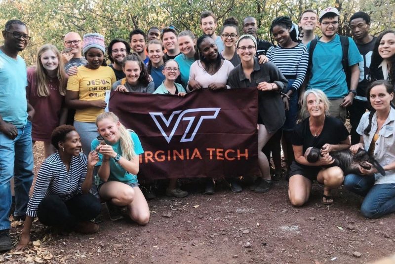 A group of people stand around a Virginia Tech flag. Two people on the right side are holding a honey badger. 