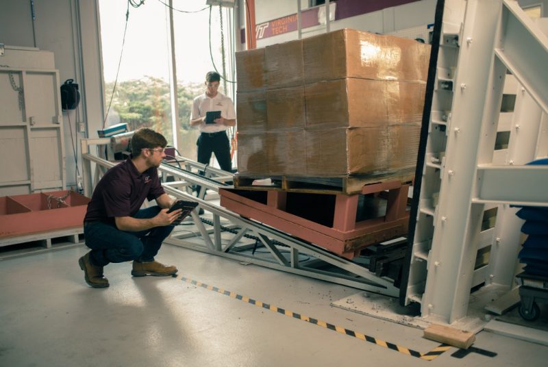 A student crouches on the ground, holding a clipboard, and looking at a machine with a slanting metal ramp supporting a shrink-wrapped stack of cardboard boxes. 