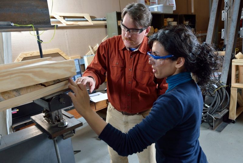 A male professor and female student study and touch the ends of stack of boards that are being held in a vice. 