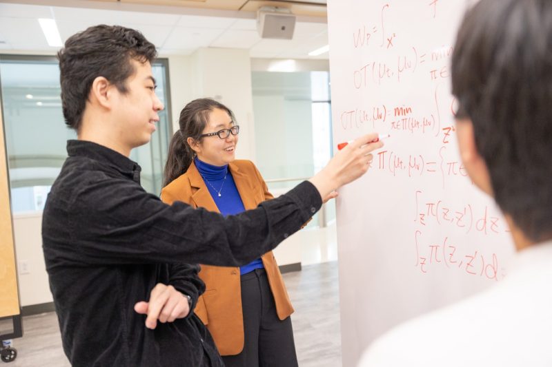 Ruoxi Jia and two students work on equations together on a large white board.