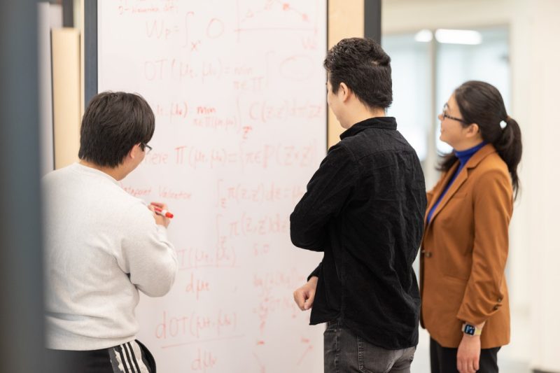 Students and professor work on problem on white board together.