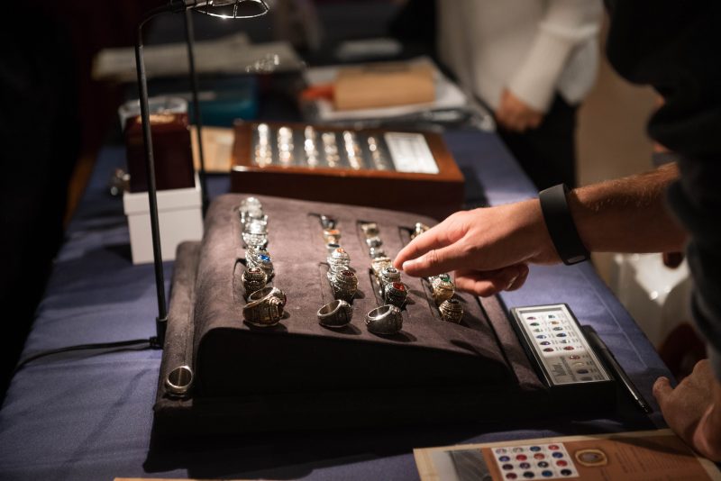 A person's hand hovers over a large display of silver and gold Virginia Tech class rings that have an assortment of multicolored stones in the middle of them. 