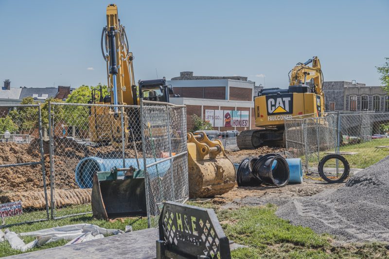 Several large yellow excavators and big blue pipes in surrounded by fencing with grey Hokie Stone Torgersen Bridge in the background