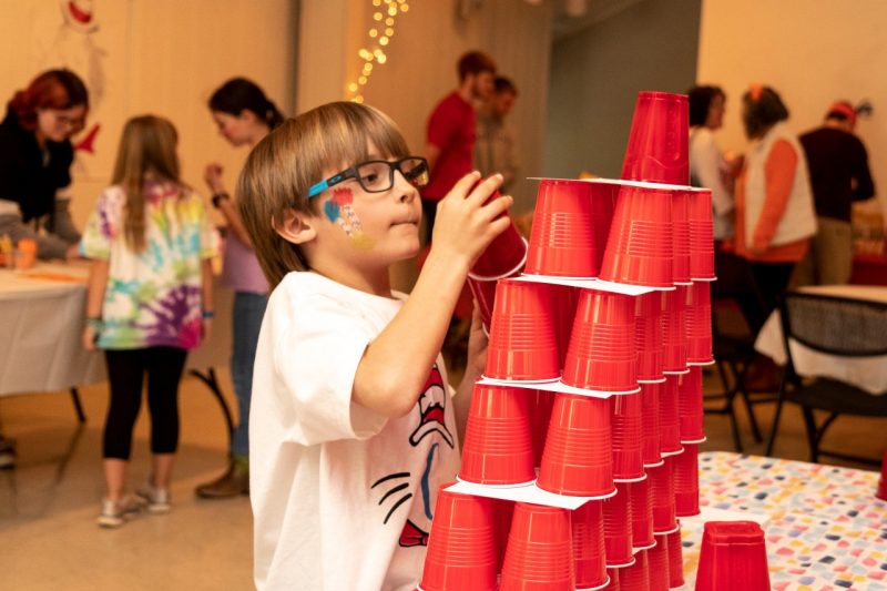 A young boy looks up at the top of a tower of red cups as he prepares to add another layer.