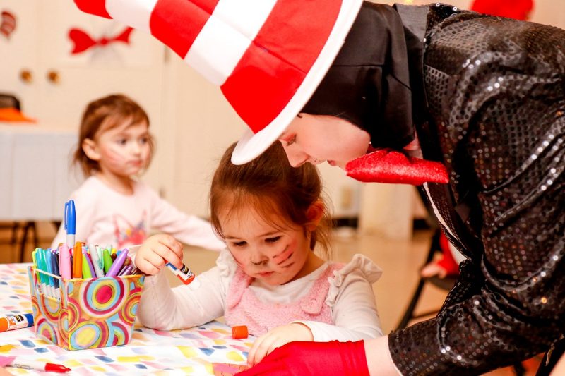 A young girl uses a glue stick with the help of a volunteer dressed in black sequence jacket, oversized bowtie, and red and white top hat.
