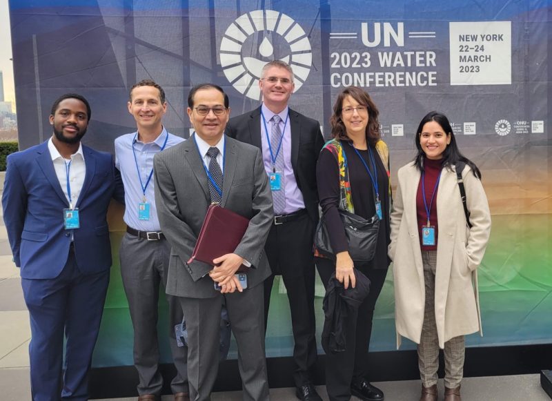 Six scholars stand in front of a display banner for the UN 2023 Water Conference. 