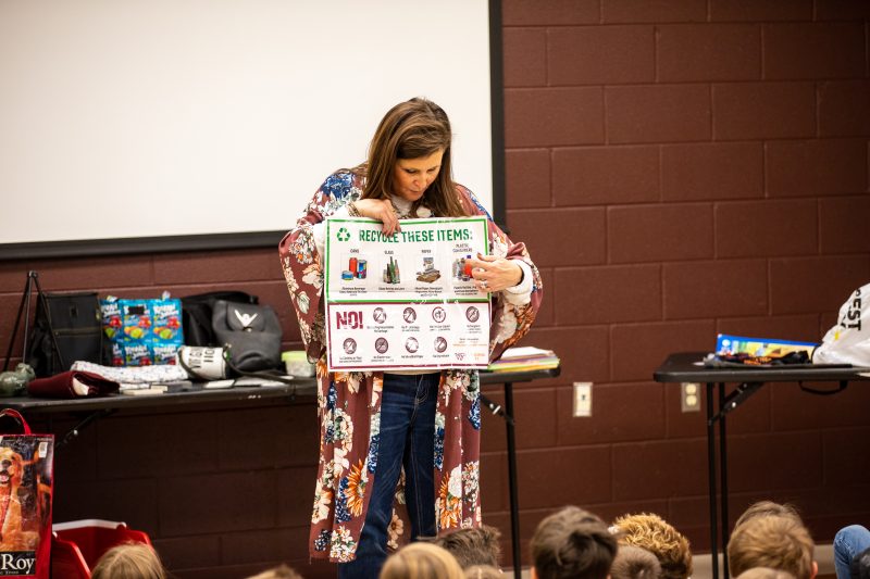 A person wearing a maroon and floral cardigan and jeans holding up a do and don't recycle poster with items printed on it that can and cannot be recycled. They are standing at the front of a classroom surrounded by many children.