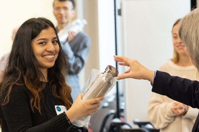 A young woman in a black shirt accepts a water bottle and smiles at the camera