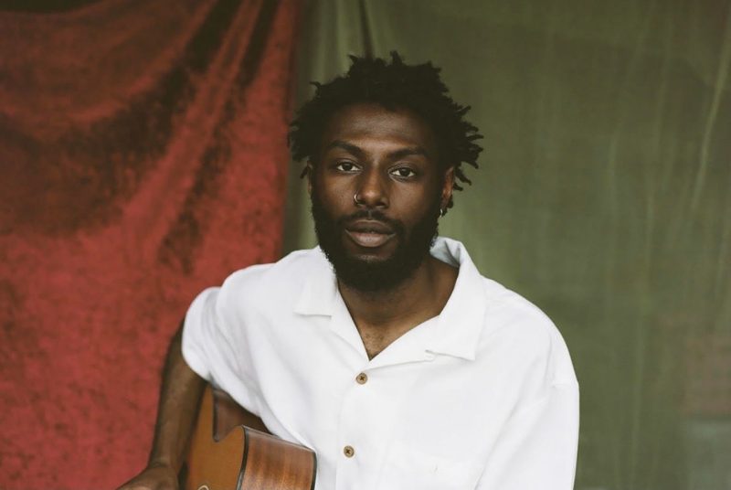 Sonny Miles is a Black man with short, twisted hair, a nose ring, and a short beard. He is wearing a white button-up shirt and holding a guitar.