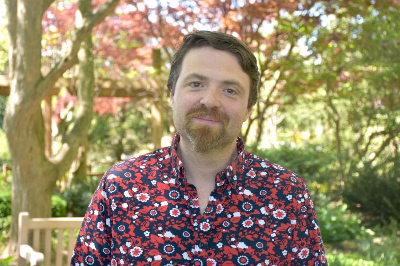 A man with brown hair and a goatee wears a flower patterned shirt  as he stands in a garden.