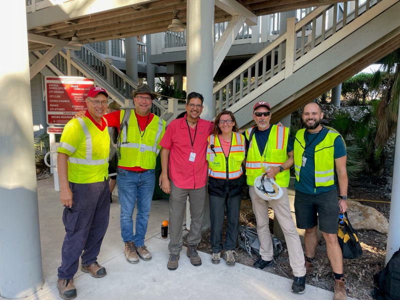 A group of Hokie alumni, all wearing work clothes and high-visibility vests, poses in front of a set of metal stairs