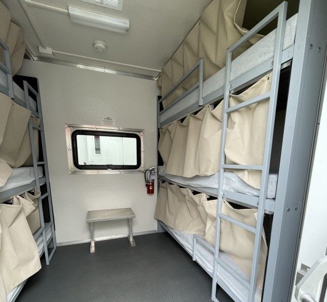 The interior of a sleeper truck. Beds wiht simple white linens are stacked three high on each side of the room with a ladder providing access to each. Each bed also has a long beige curtain that allows for some privacy.