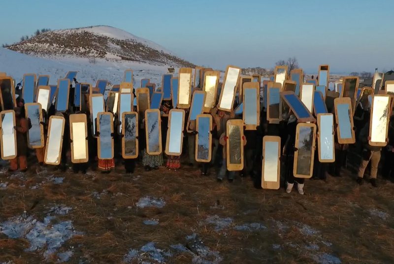 A large group of people each hold up full-length mirrors while standing outside on snowy ground, a snow-capped mountain behind them.