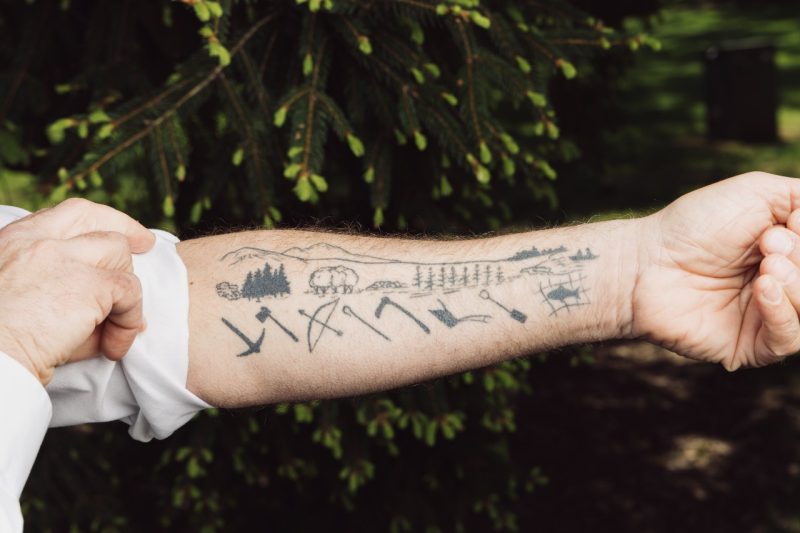 John Provo shows off the tattoo on his forearm, which has simple images of tools below a backdrop of trees, mountains, and seas.