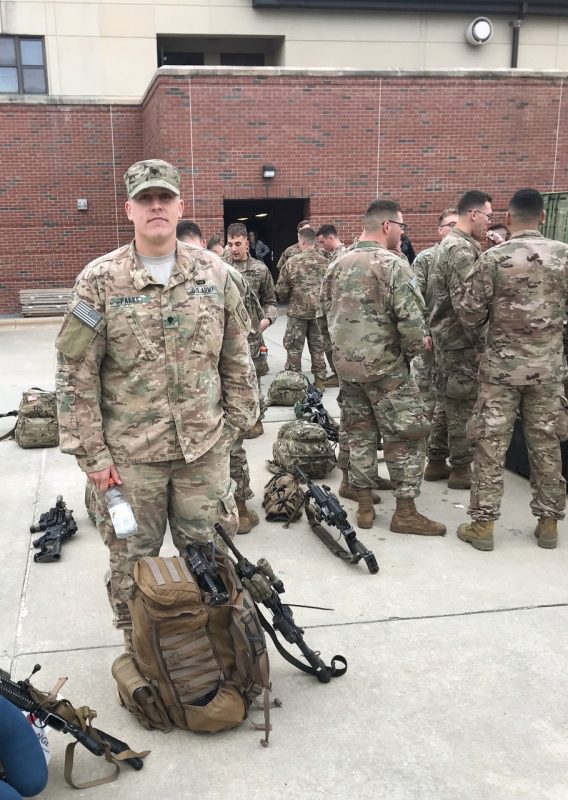 Jeff Parks stands in Army fatigues near a khaki backpack of military gear and a rifle while other men in Army fatigues mingle in the background.