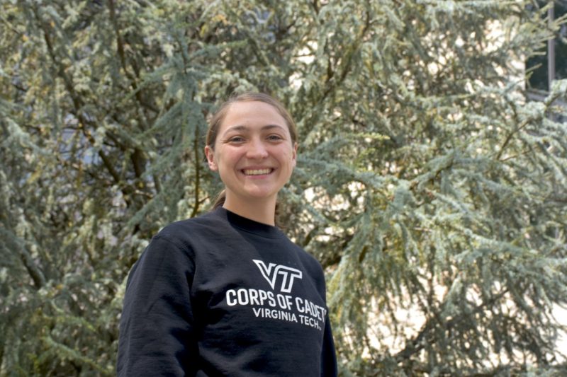 A young woman wearing a dark sweatshirt stands near a large pine tree.