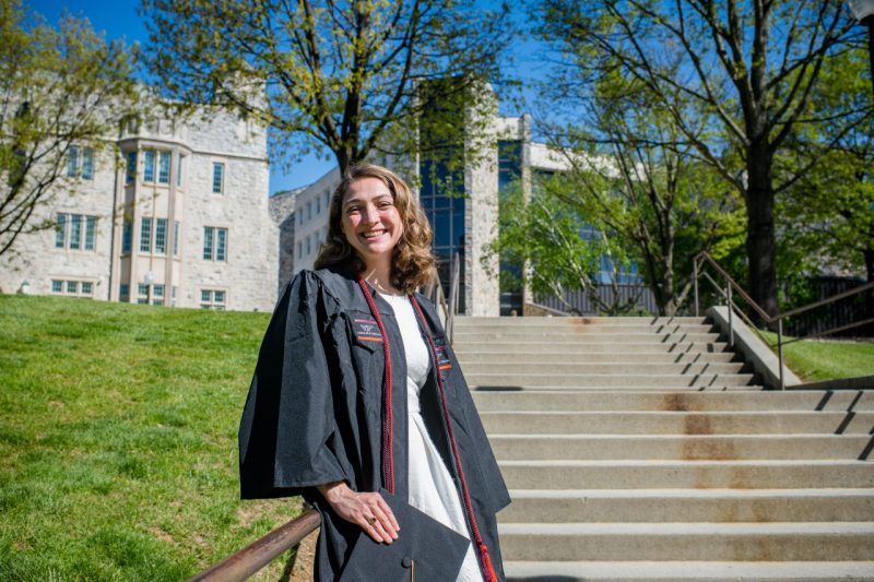 A young woman in a white dress under a black graduation gown smiles as she stands near McBryde Hall.