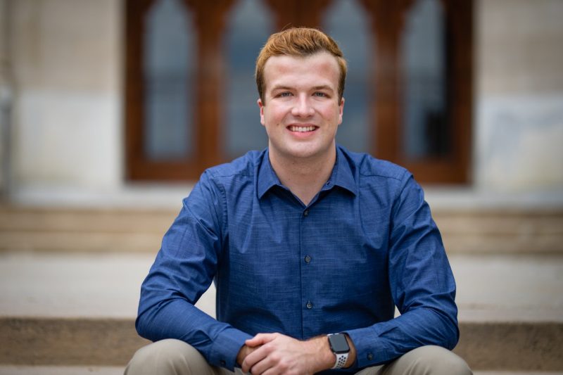 A young man with red hair and a sharp blue dress shirt sits on the stairs of Burruss Hall.