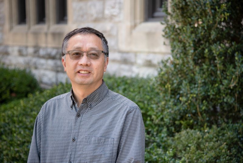 A man with salt and pepper hair, glasses, and a checkered shirt poses near a Hokie Stone building.