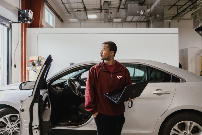 A student holds a computer in front of a car in a garage