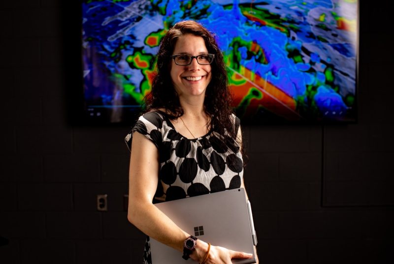 A person holding a laptop stands in front of a screen showing a meteorological map.