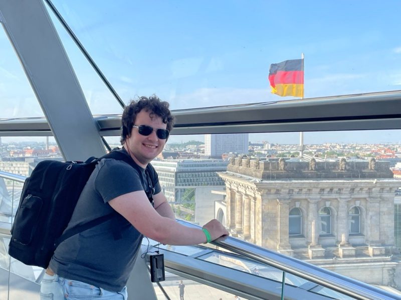student smiles at camera in building overlooking the city in Germany with the German flag in the background.